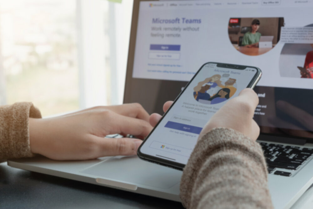 Hands holding an iPhone while on a laptop looking at Microsoft Teams