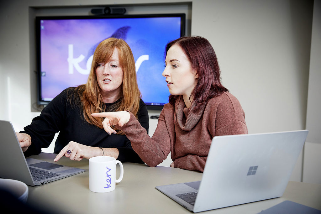 Two women pointing to a laptop with a Kerv mug in between them