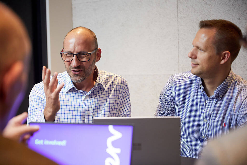 Two men sitting at laptop in office talking to a third man